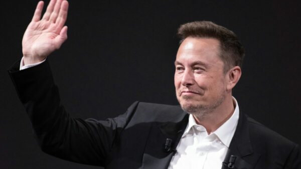 Elon Musk smirking and waving, wearing a smart suit against a black background. Tesla has cut its entire marketing team, which included a group of 40 advertising and marketing staff, just months after it was formed.