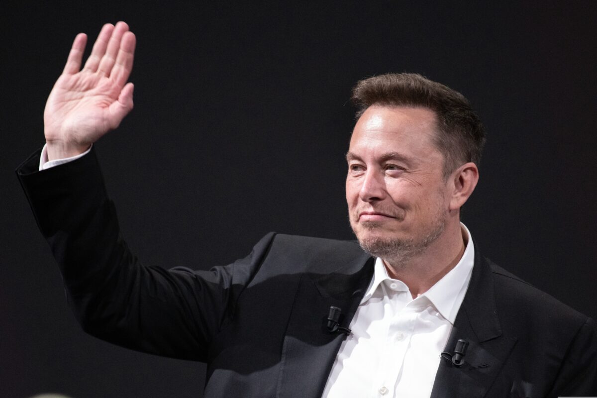 Elon Musk smirking and waving, wearing a smart suit against a black background. Tesla has cut its entire marketing team, which included a group of 40 advertising and marketing staff, just months after it was formed.