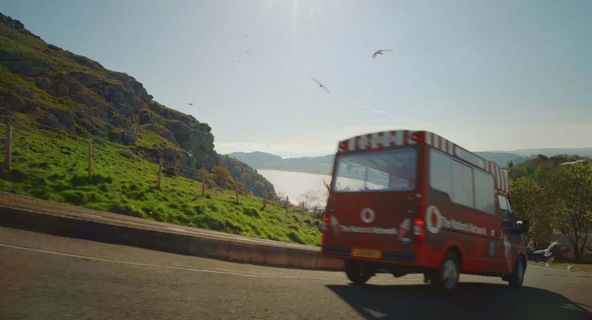 A vodafone brand ice cream van heading through the British countryside. The sea peaks out from between the hills. The van looks vintage, evoking nostalgic memories.